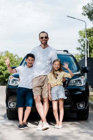Happy father in sunglasses standing with kids waving hands near carの写真素材