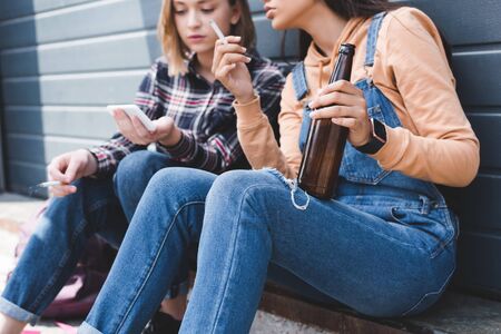 Selective focus of pretty friends smoking cigarettes, holding beer, sitting and looking at smartphoneの写真素材