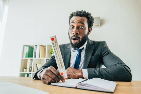 Shocked African American businessman looking at thermometer while suffering from heat in officeの写真素材