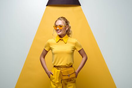 cheerful blonde woman in sunglasses standing with hands in pockets on white and yellowの写真素材
