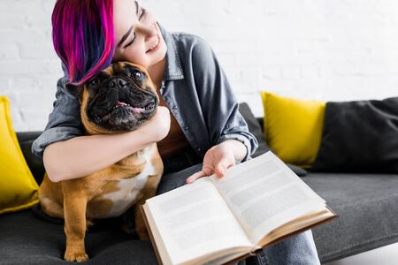 girl with colorful hair hugging bulldog, holding book and sitting on sofaの写真素材