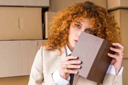 redhead woman covering face with book near cardboard boxes