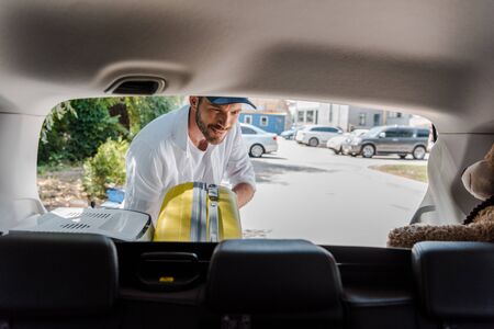 selective focus of happy man in cap putting travel bag in car trunkの写真素材