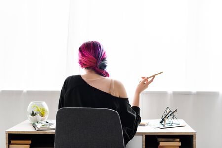back view of hipster girl holding pencil and sitting at table in living room