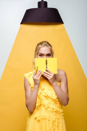 blonde young woman covering face with notebook on white and yellowの写真素材