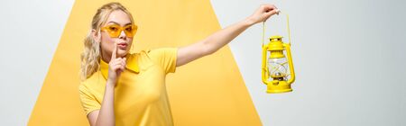 panoramic shot of attractive blonde woman in sunglasses gesturing and holding vintage lamp on white and yellowの写真素材