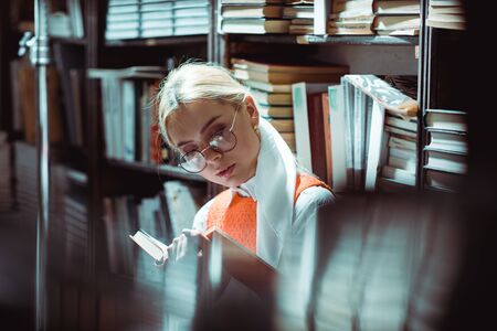 beautiful and blonde woman in glasses reading book in library