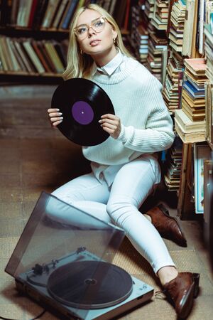 pretty and blonde woman sitting on floor and holding vinyl in libraryの写真素材