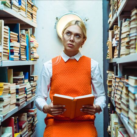 beautiful and pensive woman in orange dress holding book and looking up in library