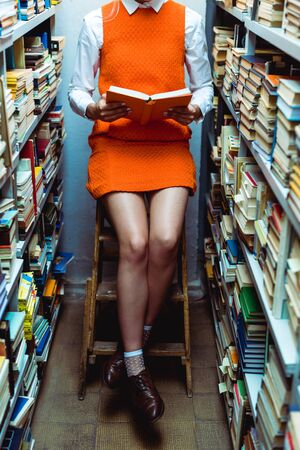 cropped view of woman in orange dress reading book and sitiing on ladder in library