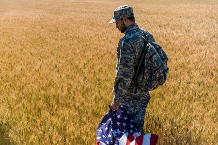 patriotic soldier in military uniform holding american flag while standing in field with wheatの写真素材