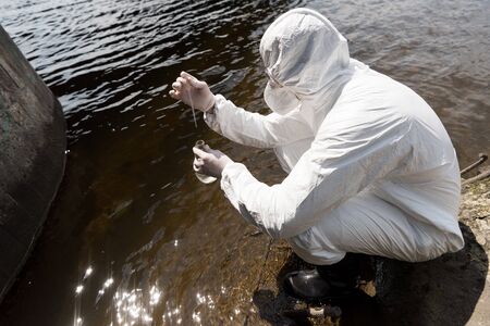water inspector in protective costume, latex gloves and respirator taking water sample at river