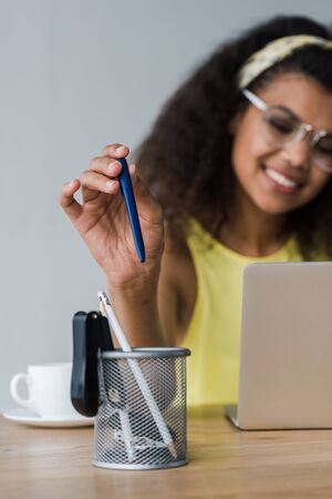 selective focus of pen holder and happy african american woman taking pen while using laptopの写真素材