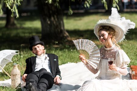 selective focus of happy victorian woman siting on blanket and holding wine glass near manの写真素材