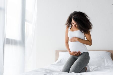 happy african american pregnant woman touching belly while sitting on bedの写真素材