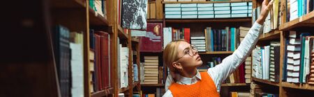 panoramic shot of pretty and blonde woman in glasses holding book in library
