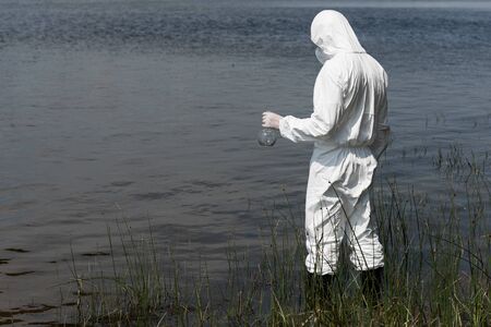 water inspector in protective costume holding flask while standing near river