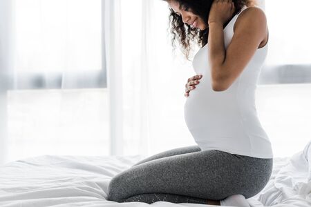 cropped view of happy curly pregnant african american woman touching belly while sitting on bedの写真素材