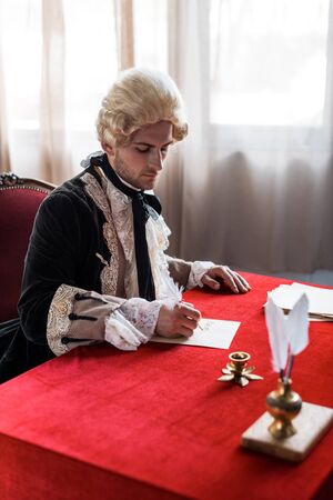 selective focus of handsome victorian man in wig holding feathers and writing letter