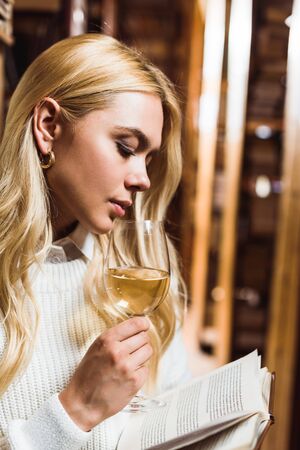 blonde and attractive woman reading book and holding wine glass in libraryの写真素材