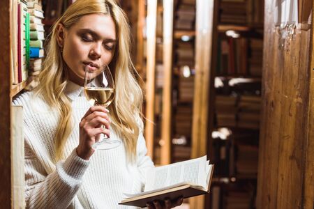 blonde woman with closed eyes holding book and wine glass in libraryの写真素材