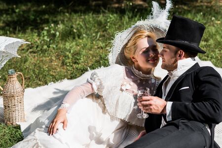 happy victorian woman siting on blanket and looking at man in hat holding wine glass