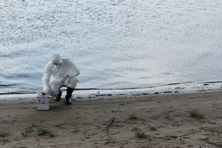 water inspector in protective costume and respirator with inspection kit sitting at river coast