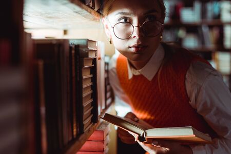 beautiful woman in glasses holding book and looking away in libraryの写真素材
