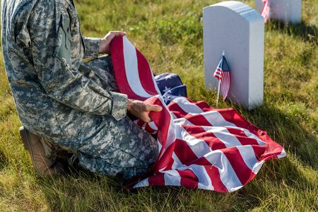 cropped view of soldier in camouflage uniform holding american flag and sitting in graveyardの写真素材