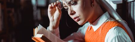 panoramic shot of beautiful woman holding glasses and reading book in libraryの写真素材