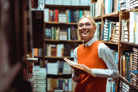 smiling and beautiful woman in orange dress holding book and looking away in libraryの写真素材
