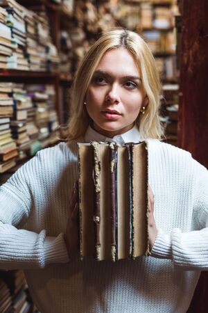blonde and pretty woman holding books and looking away in library