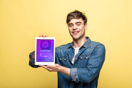 handsome smiling man in denim clothes showing digital tablet with shopping app, isolated on yellow