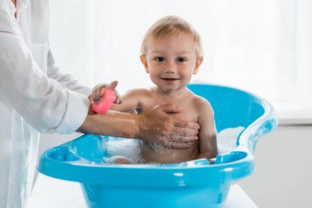 cropped view of mother washing happy toddler child with rubber toy in blue baby bathtub
