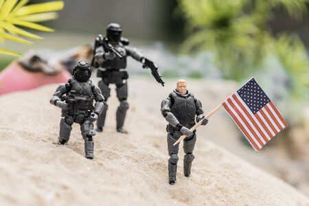 selective focus of plastic toy soldiers aiming with guns at toy man with american flag on sand hill