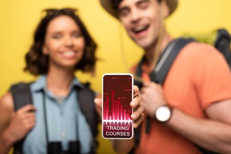 selective focus of cheerful woman showing smartphone with trading courses app while standing near...