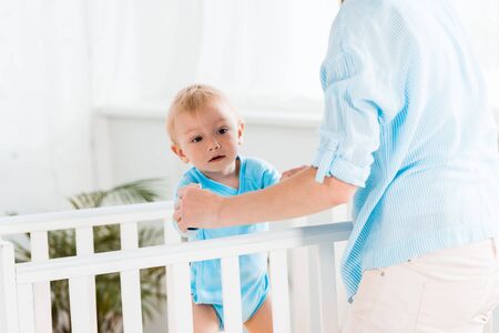 cropped view of mother standing near toddler son in baby crib