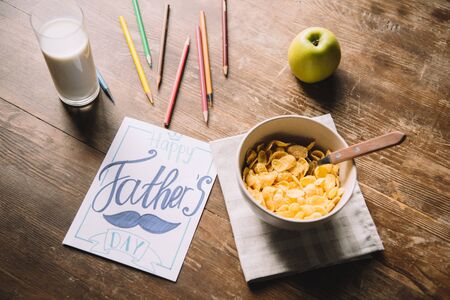 fathers day greeting card, multicolored pencils, bowl with flakes, fresh apple and glass of milk on...