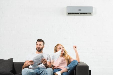 couple on couch with newspapers suffering from heat under air conditioner at homeの写真素材