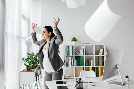 irritated pregnant woman throwing papers in air while standing near table in officeの写真素材