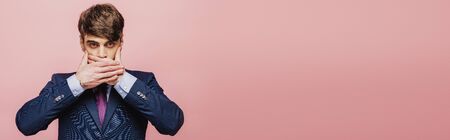panoramic shot of serious businessman holding hands on mouth while looking at camera  on pink backgroundの写真素材