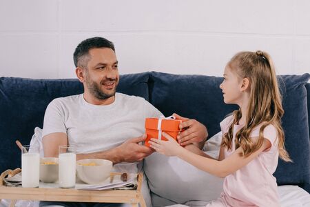 adorable daughter presenting fathers day gift box to smiling dad having breakfast in bedの写真素材
