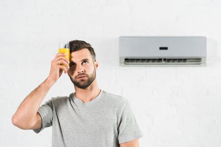 man holding orange juice near forehead and suffering from heat under air conditioner at home