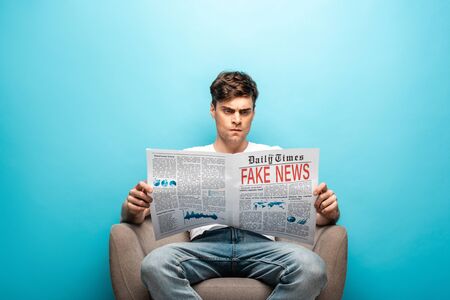 angry young man reading newspaper with fake news while sitting in armchair on blue background