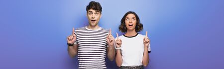 panoramic shot of cheerful man and woman looking up and pointing with fingers on blue background