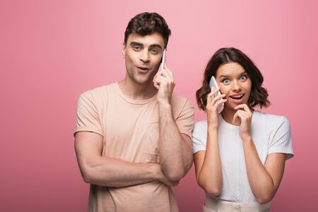 cheerful man and woman talking on smartphones while looking at camera on pink background