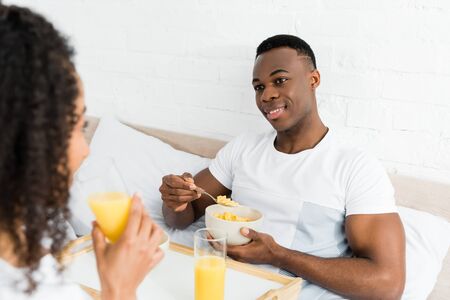 selective focus of african american man with breakfast bowl looking at girlfriend, lying in bedの写真素材