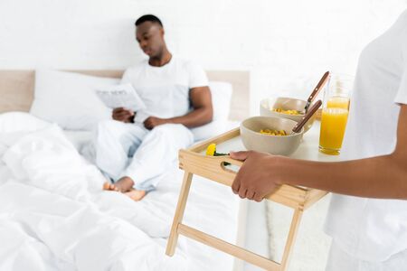selective focus of african american woman holding tray with breakfast, standing near bed in bright roomの写真素材