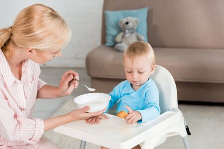 blonde mother holding bowl and feeding toddler son at home