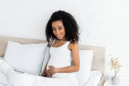pregnant african american woman looking at camera, sitting on bed in room